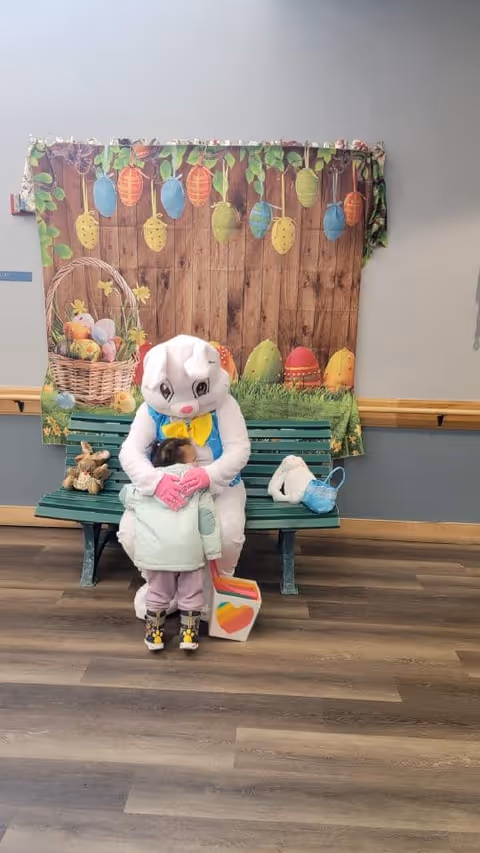 A person dressed in a white Easter Bunny costume with a yellow bow tie and blue vest sits on a green bench indoors, hugging a small child wearing a light green jacket and pink pants. Behind them is a decorative backdrop featuring colorful hanging Easter eggs and a basket filled with Easter eggs. A small stuffed bunny and a white bag with a blue mask rest on the bench beside them. The floor is wooden, and the walls are painted gray with a wooden chair rail.