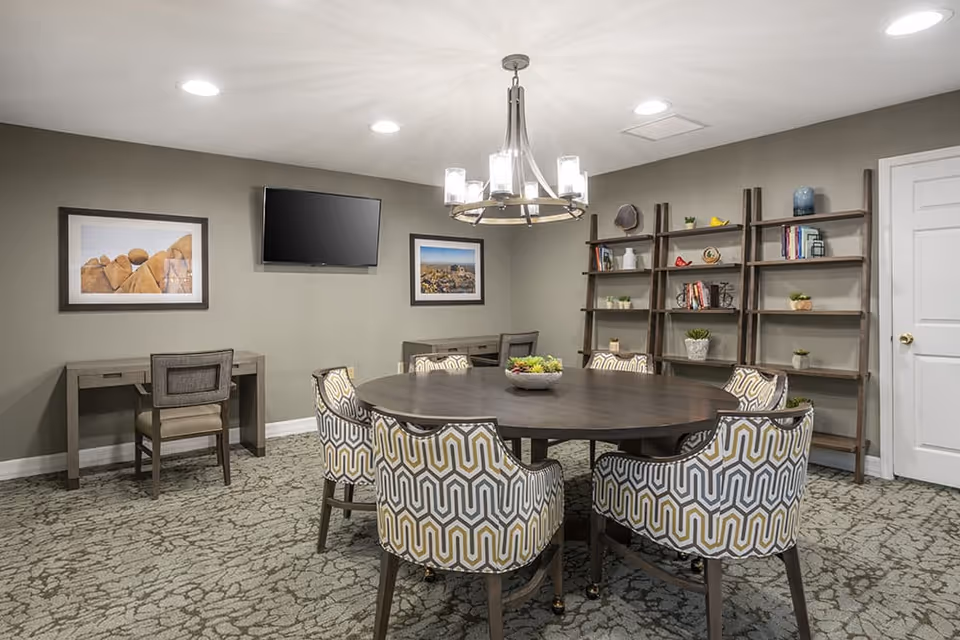 A well-lit interior room with a round wooden table surrounded by six patterned upholstered chairs. The room has a carpeted floor, two desks with chairs against the wall, a wall-mounted flat screen TV, two framed landscape photographs, and a tall wooden bookshelf with various decorative items and books. A modern chandelier hangs from the ceiling.
