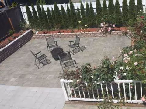 Outdoor paved patio with metal chairs around a small table, surrounded by railings and plants.