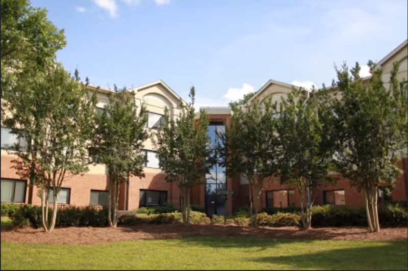Exterior view of a two-story brick and beige building with multiple windows, surrounded by neatly trimmed bushes and several tall trees in front, under a clear blue sky.