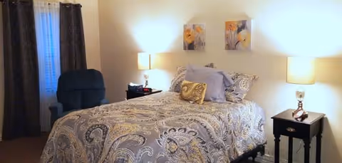 Well-lit bedroom with a patterned bed, two bedside tables with lamps, an upholstered armchair by curtained window, and artwork above the bed.