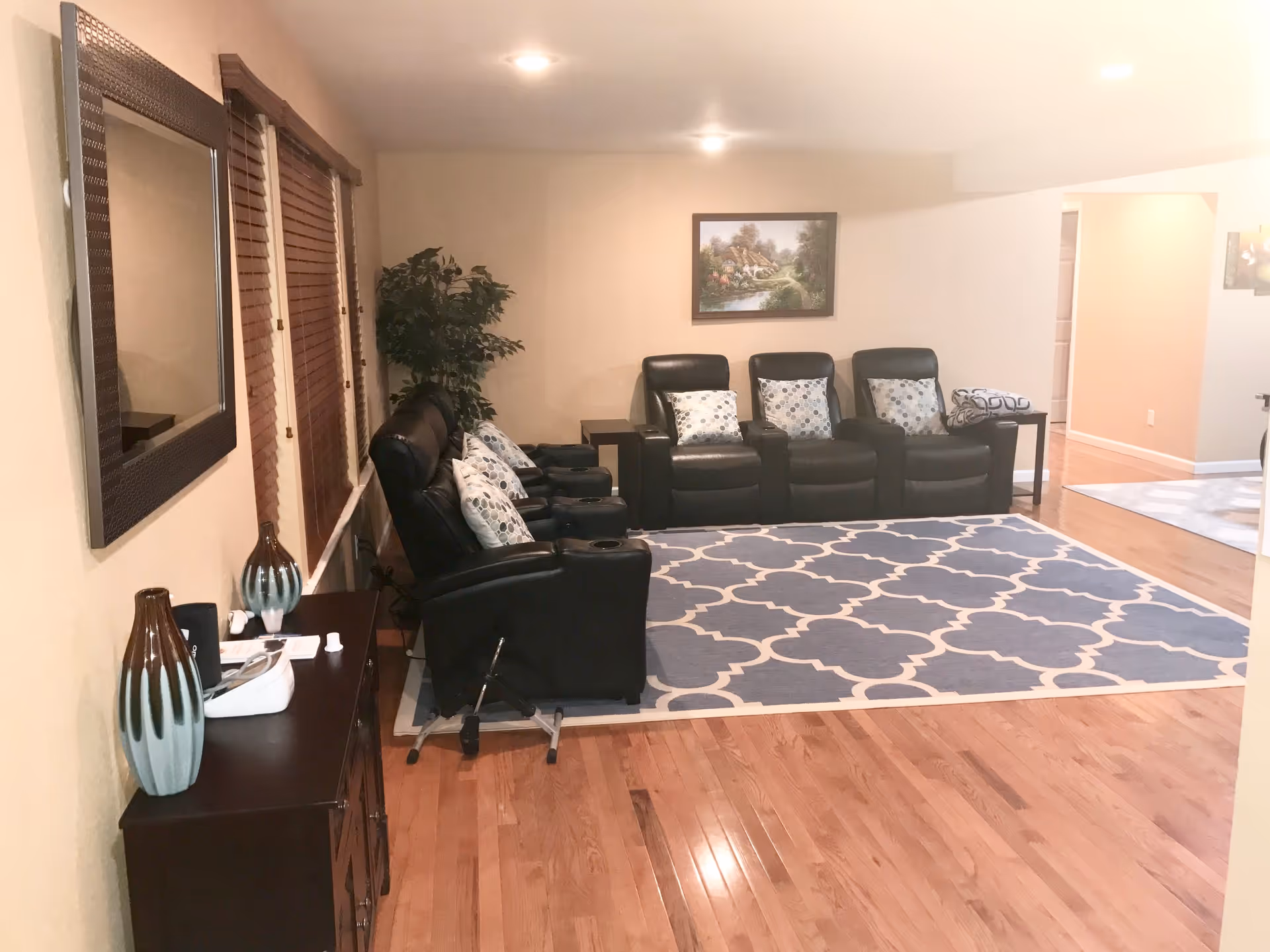 A cozy living room area with five black leather recliner chairs arranged in a row, each with patterned cushions. The floor has a large blue and white patterned rug. There is a wooden cabinet with decorative vases and a mirror on the left wall. A framed landscape painting hangs on the far wall above the chairs. Wooden blinds cover the windows on the left side, and a potted plant is placed in the corner.