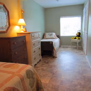 A bright bedroom with two single beds, each covered with a quilt featuring heart patterns. Between the beds are two dressers, one dark wood and one white, with a lamp and a mirror on the wall above the dark dresser. A window at the far end lets in natural light, and a chair near the window holds a stuffed animal.