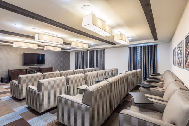 A cozy media room with multiple rows of cushioned armchairs arranged facing a wall-mounted flat screen TV. The room features modern ceiling lights, patterned curtains, and framed artwork on the walls.