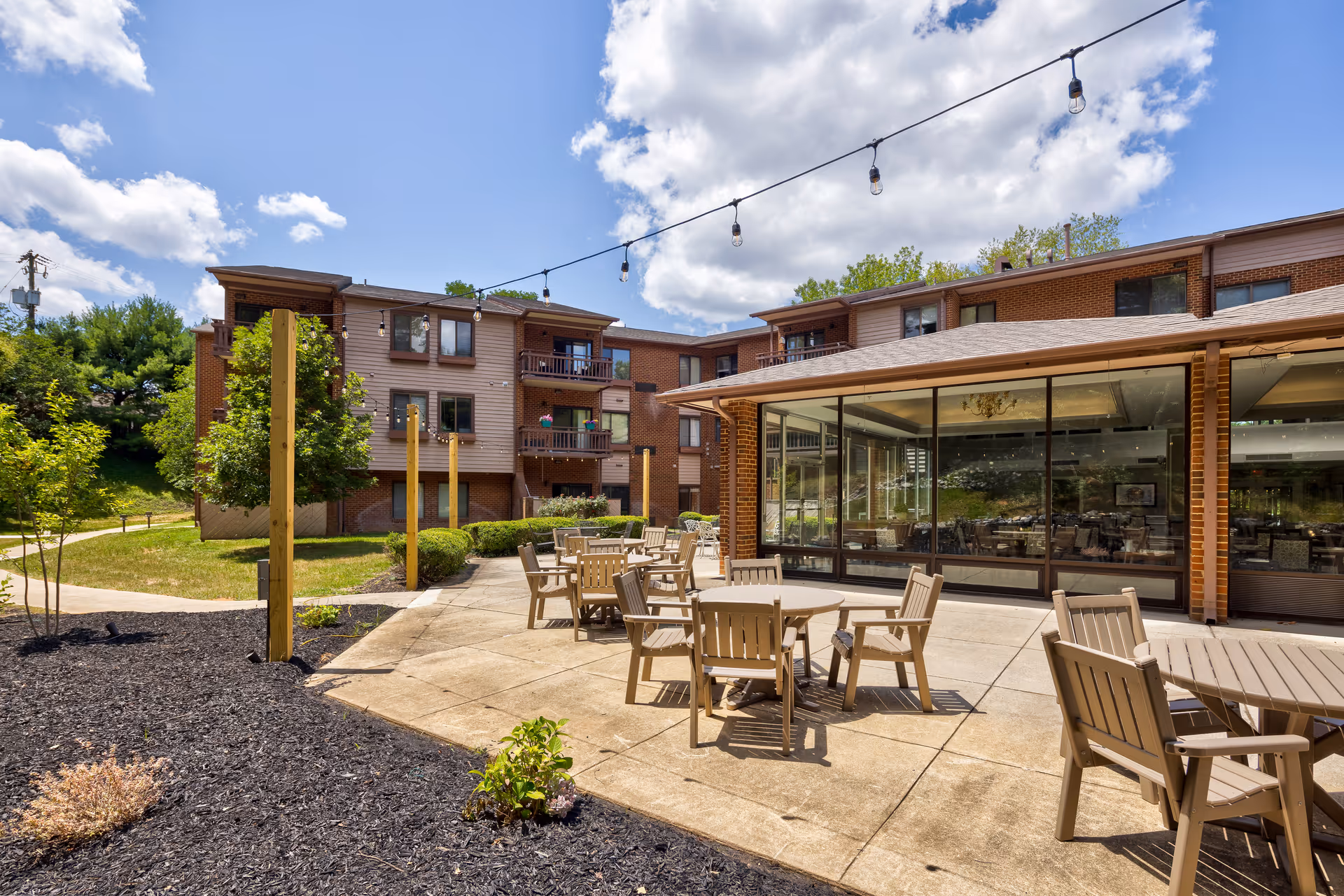 Sunny outdoor patio with round tables and chairs, string lights overhead, and a multi-story senior living building with large glass windows.