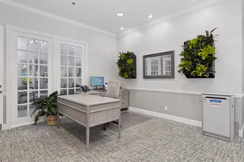 A modern office space with a light gray desk and matching chair on a patterned carpet. The desk has a computer monitor, keyboard, and phone. Behind the desk is a set of double glass doors with white frames. The walls are painted white and light gray with two green vertical plant arrangements and a rectangular mirror hanging on the wall. A small potted plant is on the floor near the doors, and a small white cabinet is positioned against the wall.