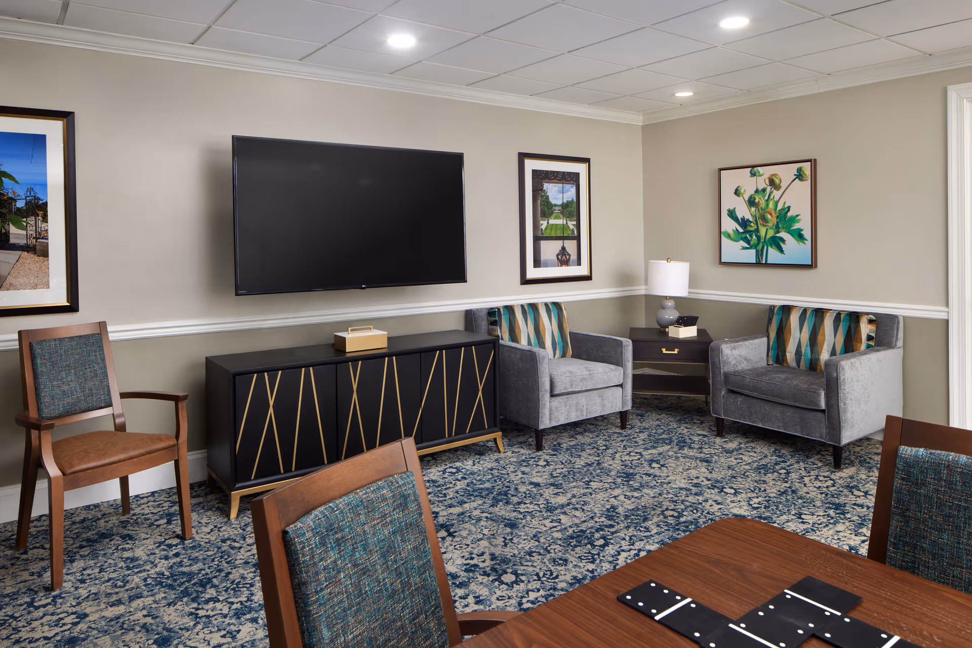A cozy living room area with two gray armchairs featuring patterned cushions, a wooden side table with a lamp, a black TV cabinet with gold accents, a wall-mounted flat-screen TV, and framed artwork on beige walls. There are also wooden chairs with teal upholstery around a table with dominoes on it, and a blue patterned carpet covering the floor.