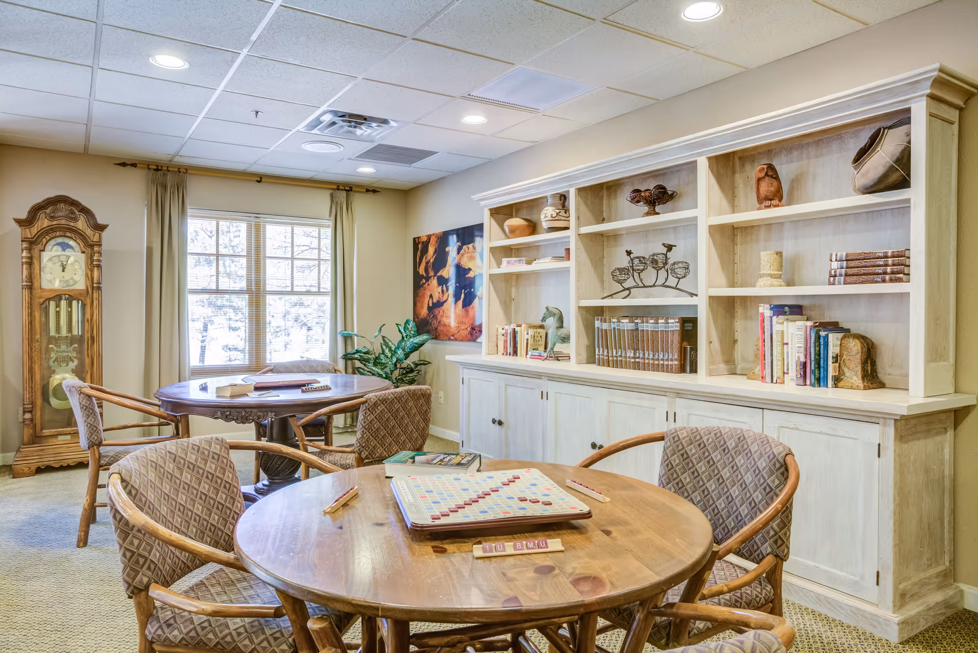 A cozy senior living community room with a round wooden table and four cushioned chairs arranged around it. A Scrabble board game is set up on the table. In the background, there is a second round table with chairs near a window with beige curtains. A tall grandfather clock stands beside the window. On the right side, a large white wooden bookshelf holds books and decorative items. The room has a carpeted floor and a ceiling with recessed lighting.