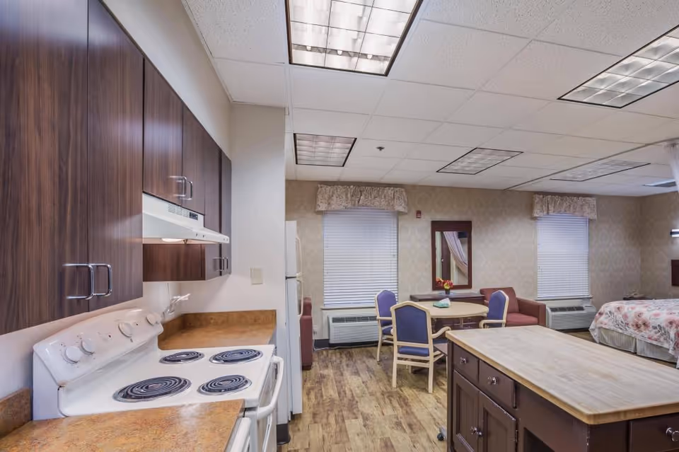 Interior view of a senior living facility room featuring a small kitchen area with a stove, oven, and wooden cabinets on the left. The room also includes a dining table with four chairs, a mirror on the wall, two windows with blinds and valances, a small couch, and a bed with floral bedding on the right. The flooring is wood-style, and the ceiling has recessed fluorescent lighting.