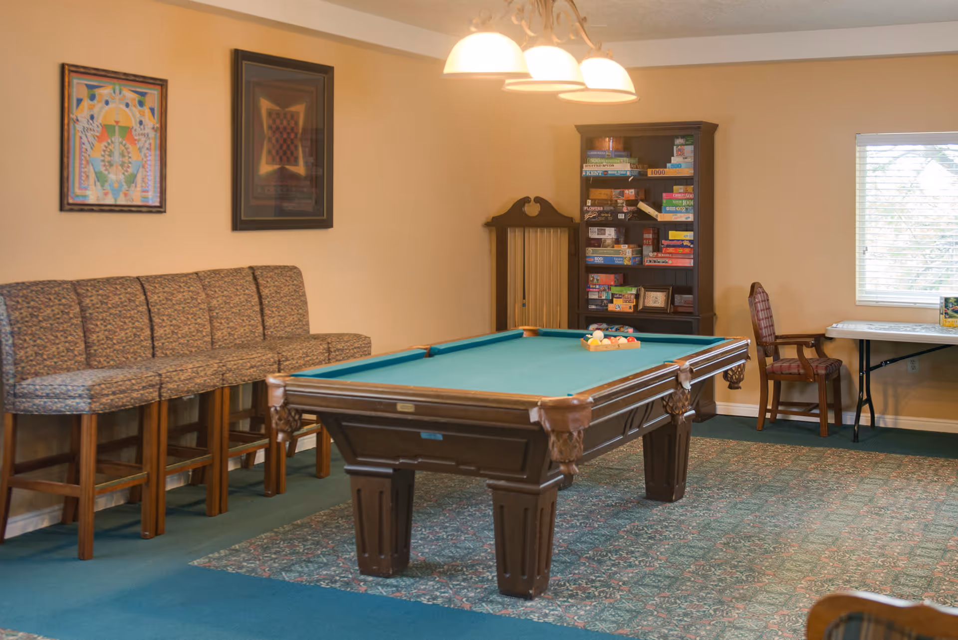 A recreational room with a pool table in the center, a row of patterned cushioned chairs along the left wall, a bookshelf filled with board games, a wooden chair, and a table near a window with blinds. The room has beige walls and a patterned carpet.