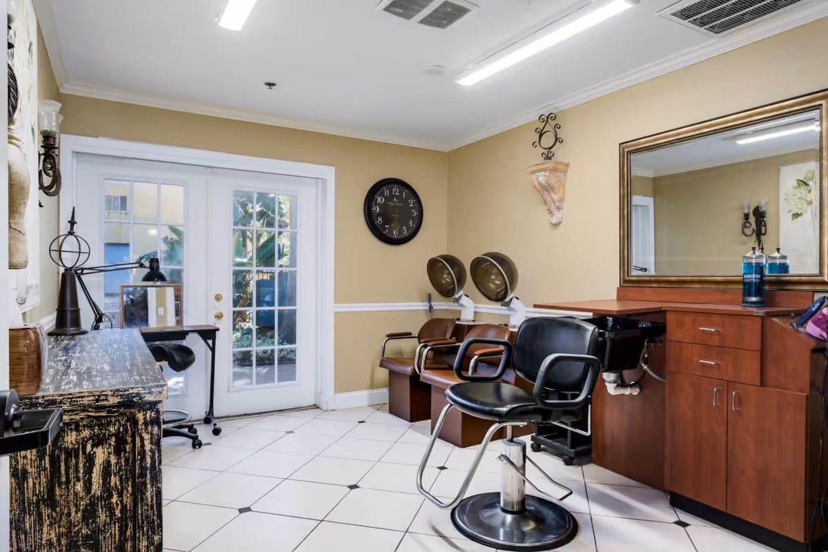 Interior view of a salon area in a senior living facility with a black salon chair in front of a wooden counter and large mirror. Two brown chairs with hair dryers are positioned against a beige wall with a black clock. A small desk with a lamp and mirror is near glass double doors leading outside.