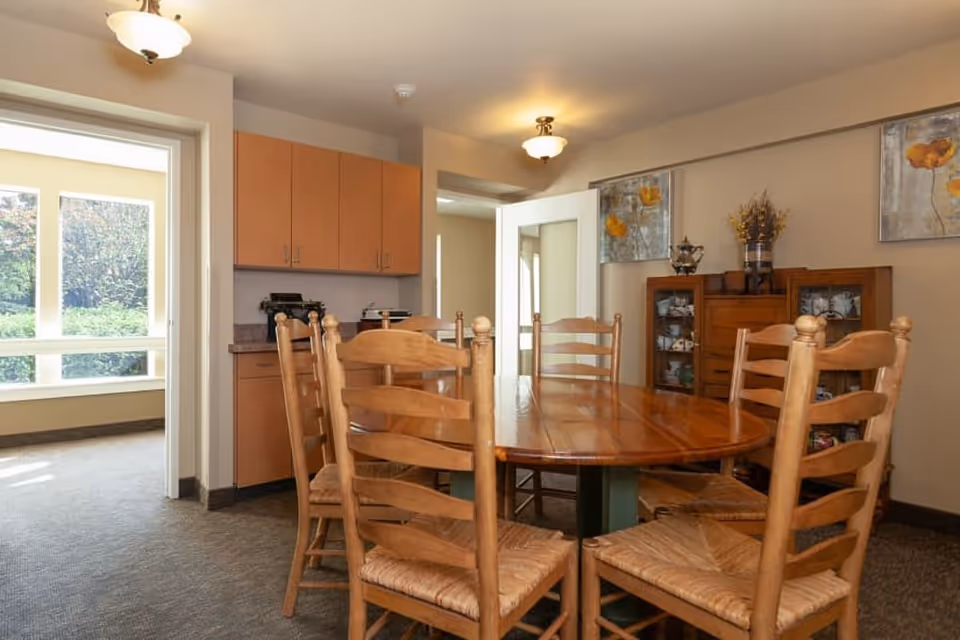 A dining area with a wooden oval table surrounded by six wooden chairs with woven seats. The room has beige walls and carpeted floor. There are two paintings with floral designs on the wall, a wooden cabinet with glass doors displaying dishes, and a small countertop with cabinets above it. A doorway leads to a bright room with large windows showing greenery outside.