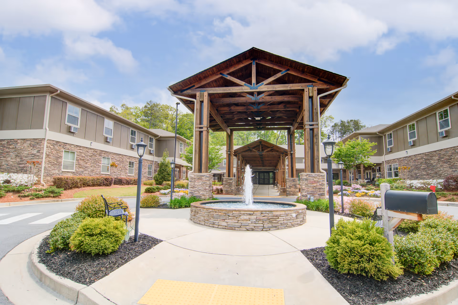 Entrance of Oaks at Towne Lake senior living facility featuring a covered wooden pavilion with stone pillars, a central water fountain, landscaped bushes and flowers, benches, and two-story buildings on either side under a partly cloudy sky.