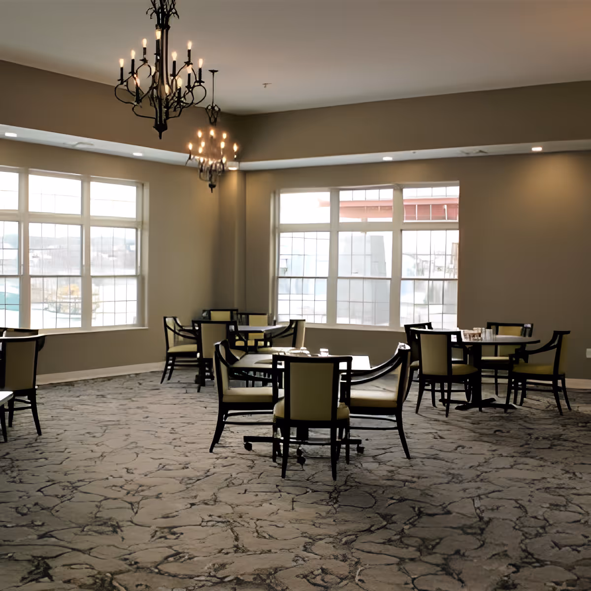 A spacious dining room with several round tables and chairs arranged neatly. Large windows allow natural light to fill the room, and two decorative chandeliers hang from the ceiling. The carpet has a textured pattern, and the walls are painted in a neutral tone.
