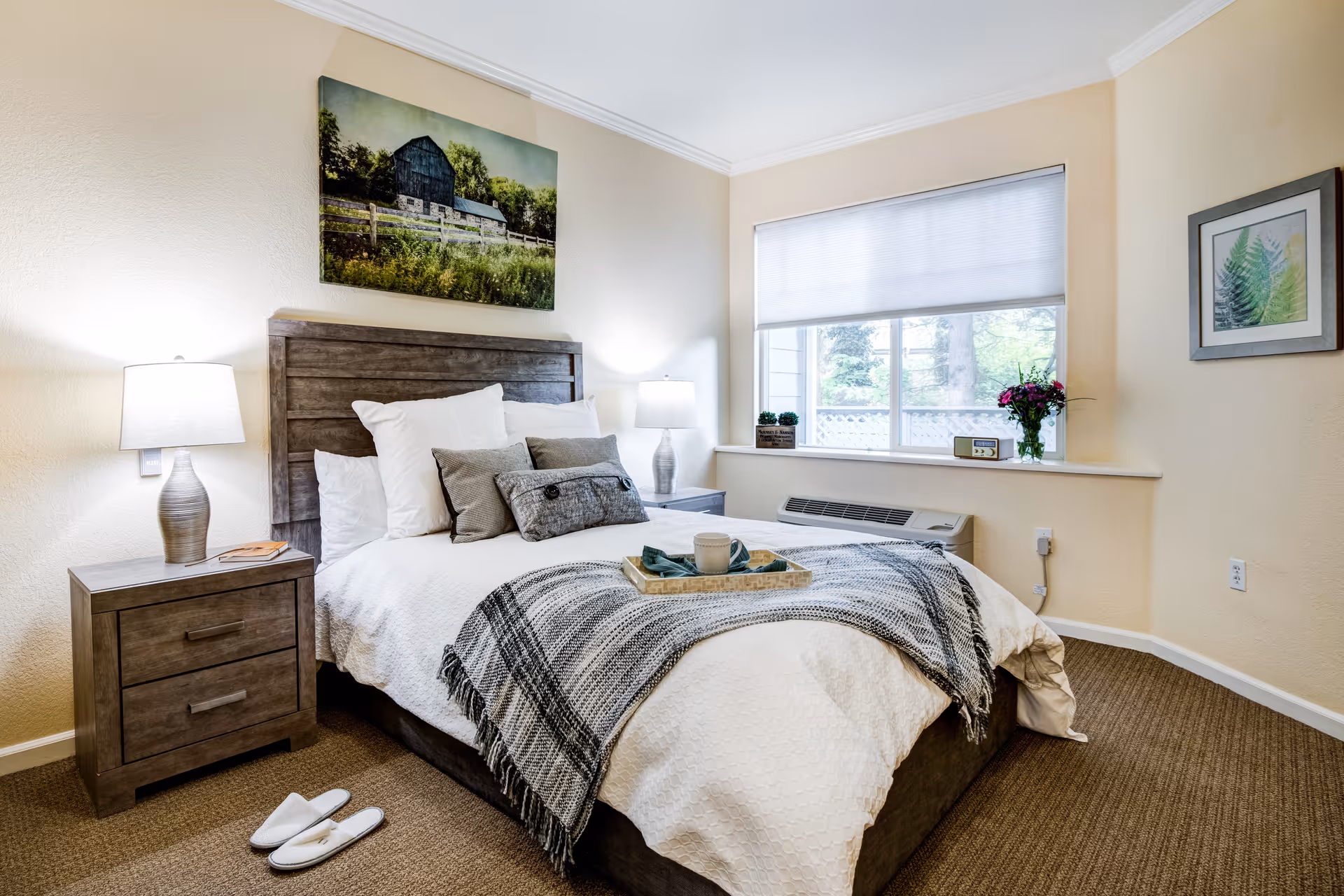 A cozy bedroom with a wooden headboard bed made with white bedding and multiple pillows. A gray and white throw blanket is draped across the foot of the bed with a tray holding a cup and napkin. There are two matching wooden nightstands with lamps on each side of the bed. A pair of white slippers is placed on the carpeted floor next to one nightstand. The room has beige walls with a large window covered by a white blind, a framed picture of a barn above the bed, and a framed botanical print on the adjacent wall. A small vase of flowers and a clock are on the window sill.