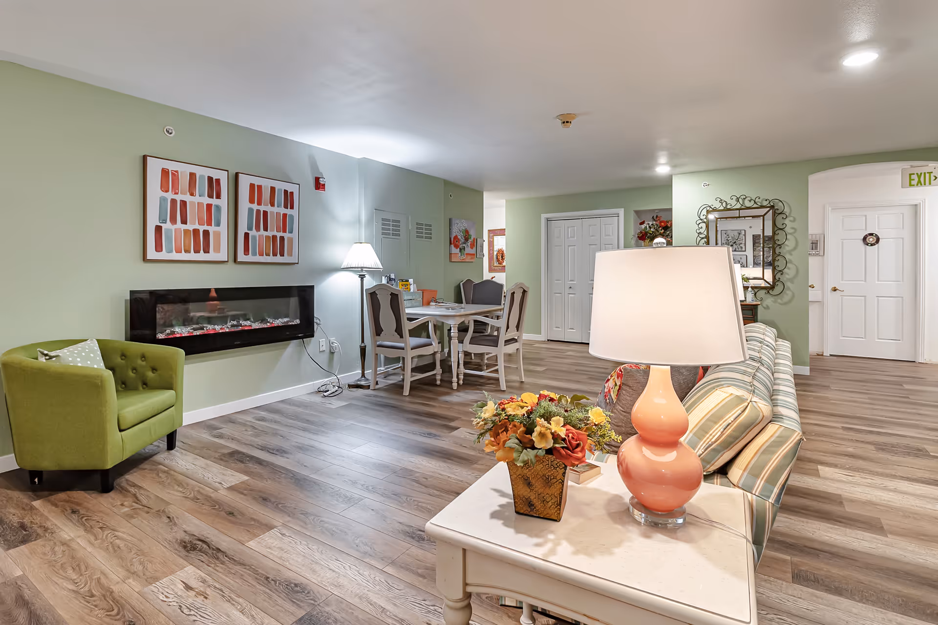 A cozy senior living common area with light green walls and wood flooring. The room features a green armchair with a polka dot pillow, a wall-mounted electric fireplace beneath two abstract art pieces, a dining table with four chairs, and a striped sofa with a side table holding a pink lamp and a floral arrangement. A large mirror and decorative items are visible in the background, along with white doors and an exit sign.