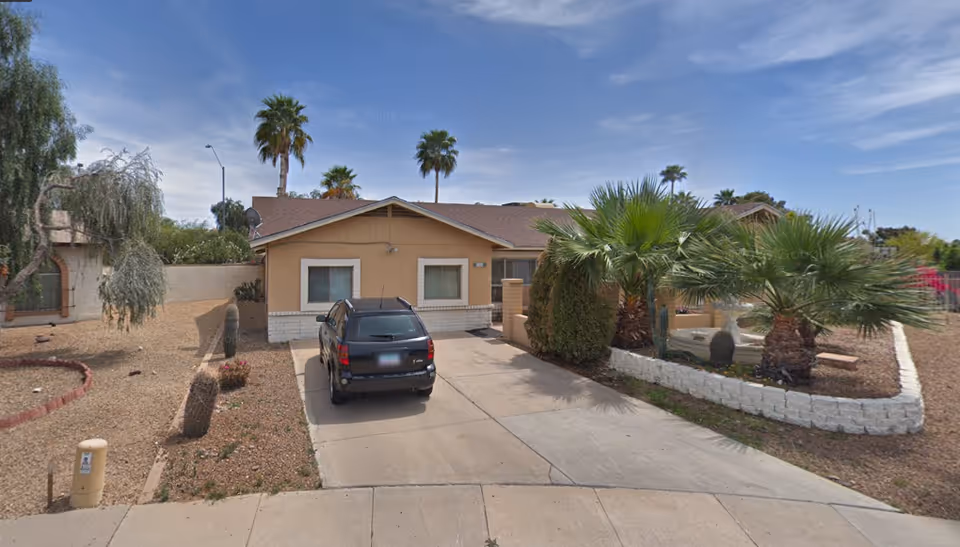 Single-story house with a beige exterior and a brown roof, a black SUV parked in the driveway, surrounded by desert landscaping including palm trees, cacti, and gravel. The sky is clear with some clouds.
