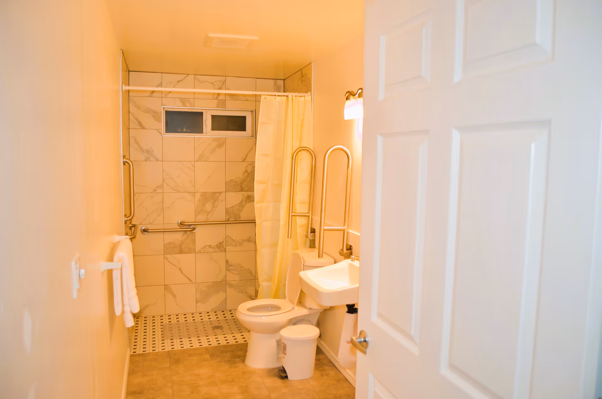 A clean, accessible bathroom with a white toilet, a small white sink, and a walk-in shower with marble-patterned tiles and a yellow shower curtain. There are grab bars near the toilet and inside the shower for support. A white towel hangs on a towel rack on the left wall, and a small white trash can is placed near the sink. The bathroom door is partially open.