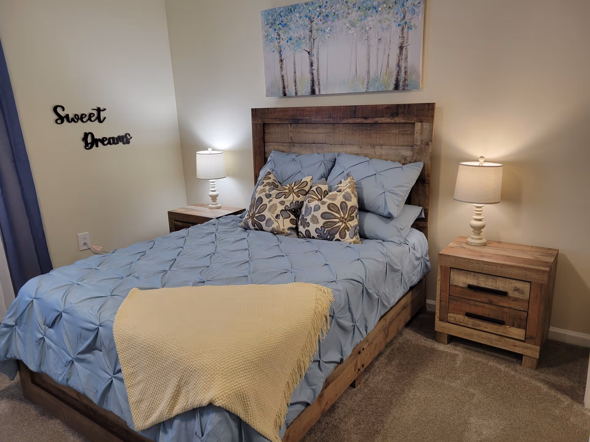 A cozy bedroom with a wooden bed frame and headboard, made up with light blue bedding, two floral patterned pillows, and a yellow throw blanket at the foot of the bed. There are two matching wooden nightstands on either side of the bed, each with a white lamp. On the wall above the bed is a painting of trees, and on the adjacent wall, black decorative letters spell out 'Sweet Dreams'. The room has beige carpet and light-colored walls.