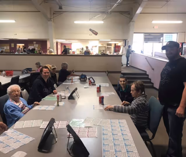 A group of elderly people and a few adults sitting around a large table playing bingo in a well-lit room with a counter and other people in the background.