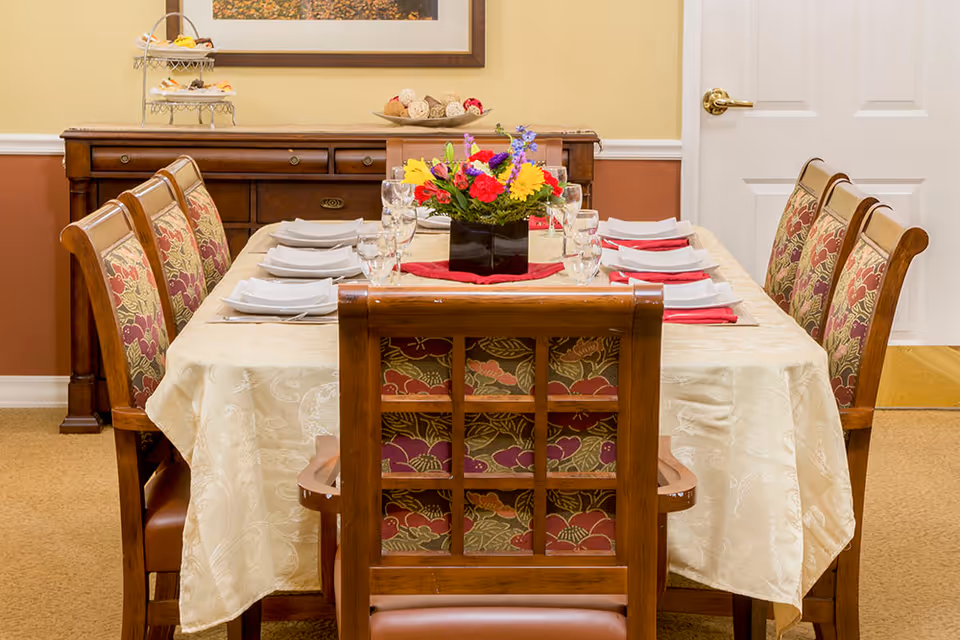 A dining room with a rectangular table covered with a cream-colored tablecloth. The table is set with white plates, folded napkins, and glassware for eight people. A colorful floral centerpiece is placed in the middle of the table. Surrounding the table are wooden chairs with floral-patterned upholstery. In the background, there is a wooden sideboard with decorative items and a framed picture on the wall.