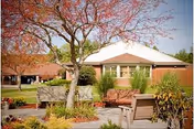 Outdoor seating area with wooden benches and chairs surrounded by plants and trees with autumn foliage, set in front of a single-story building with a white roof under a clear sky.