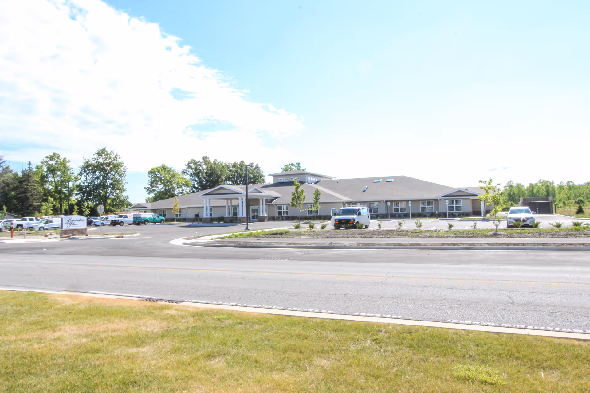 Exterior view of Lincolnshire Place - Memory Care & Assisted Living building with a parking lot in front, several cars parked, and a sign near the entrance. The building is single-story with a pitched roof and surrounded by trees and greenery under a partly cloudy sky.