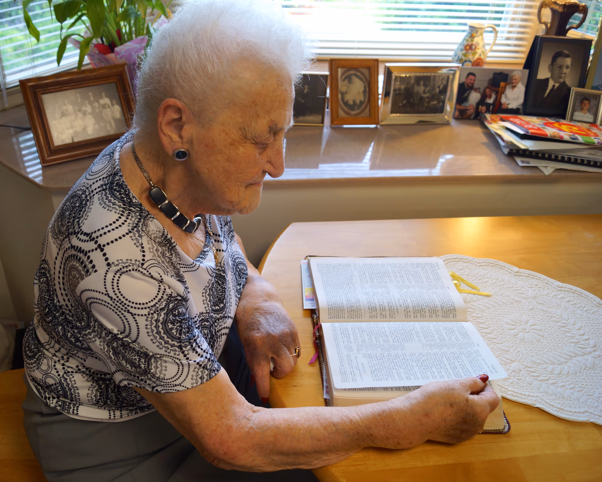 An elderly woman with white hair is sitting at a wooden table reading an open book. Behind her on a windowsill are several framed photographs and a decorative pitcher. The room is well-lit with natural light coming through the window blinds.