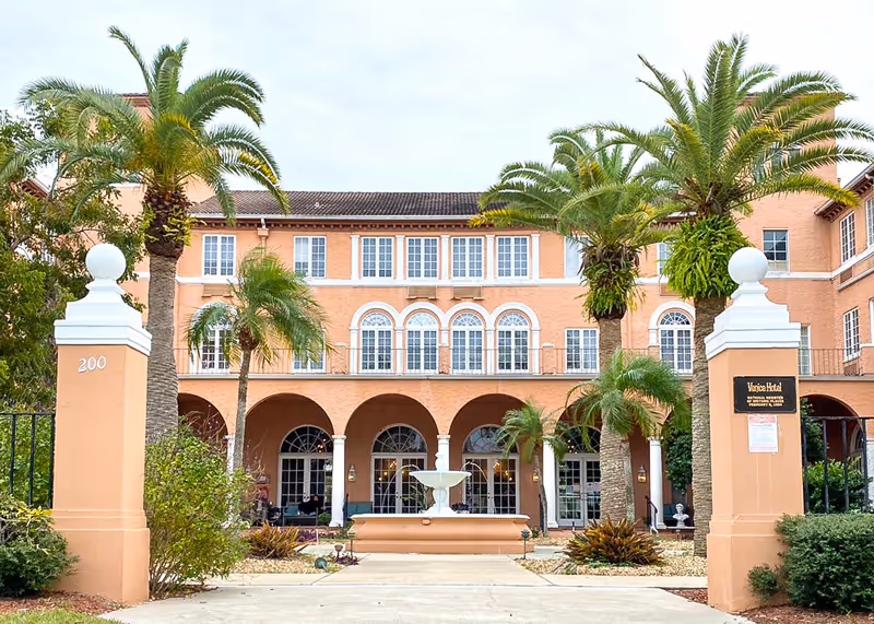 Front exterior view of a large peach-colored building with arched windows and doorways, palm trees, and a central water fountain in front. Two pillars mark the entrance with the number 200 on the left pillar and a sign on the right pillar reading 'Venice Hotel'.