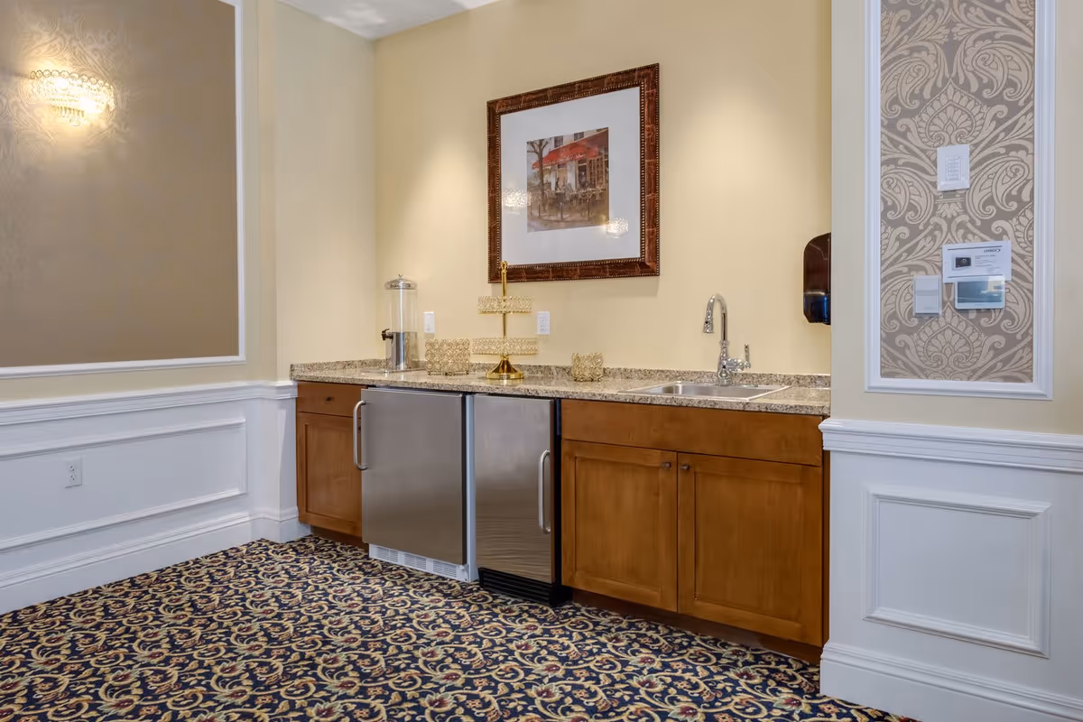 A small kitchenette with granite countertops, stainless mini-fridges, a sink, wooden cabinets, patterned carpet, and framed artwork on the wall.