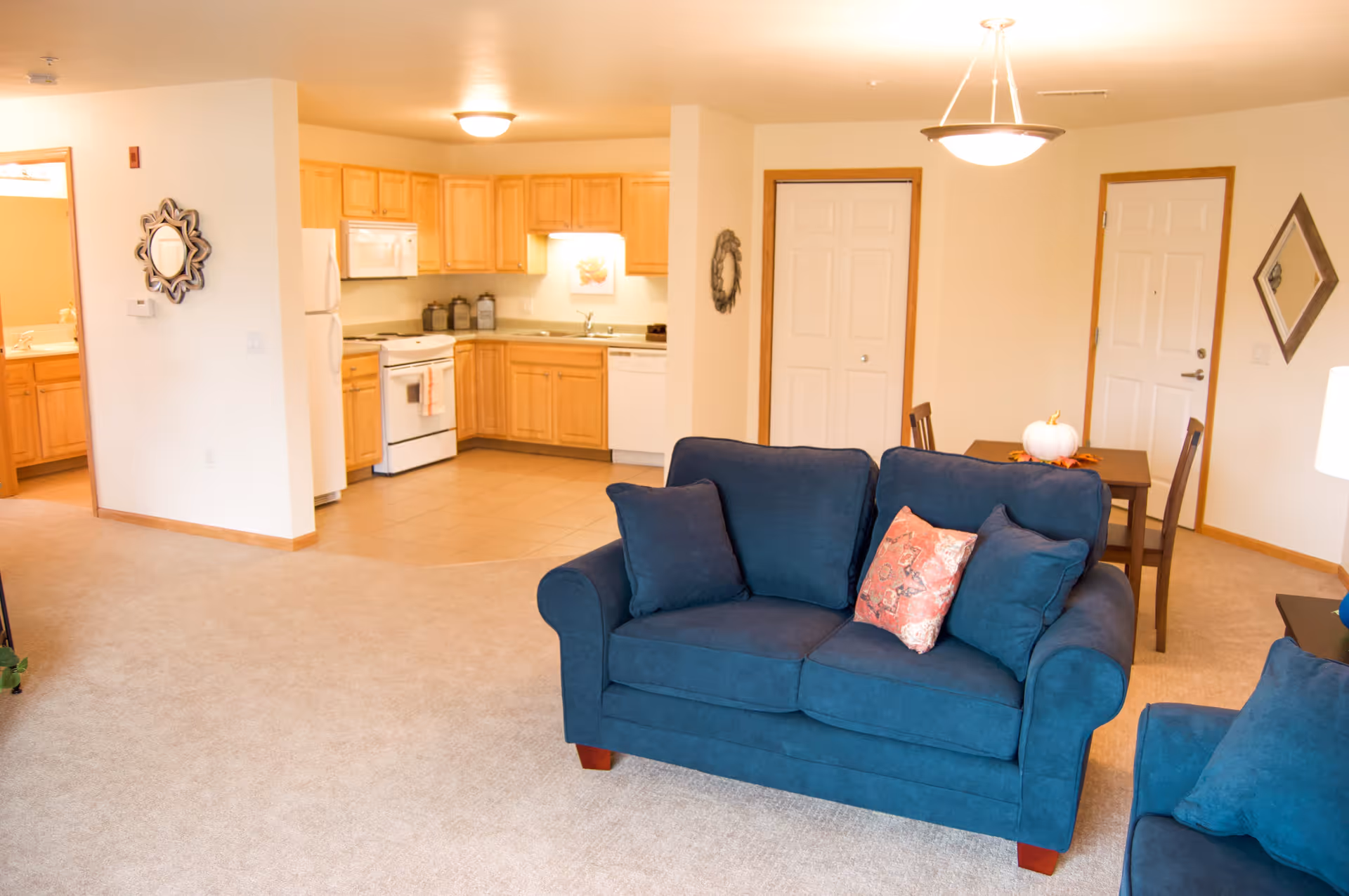 Interior view of a senior living apartment featuring a living room with blue upholstered sofas and a pink patterned pillow, an adjacent dining area with a small table and chairs, and a kitchen with wooden cabinets, white appliances, and tiled flooring. The walls are light-colored with decorative mirrors and a door leading to another room.