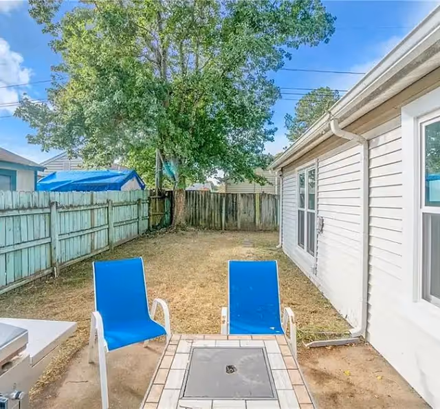 Small fenced backyard with two blue patio chairs facing a tiled fire pit, a large tree, and the side of a house.