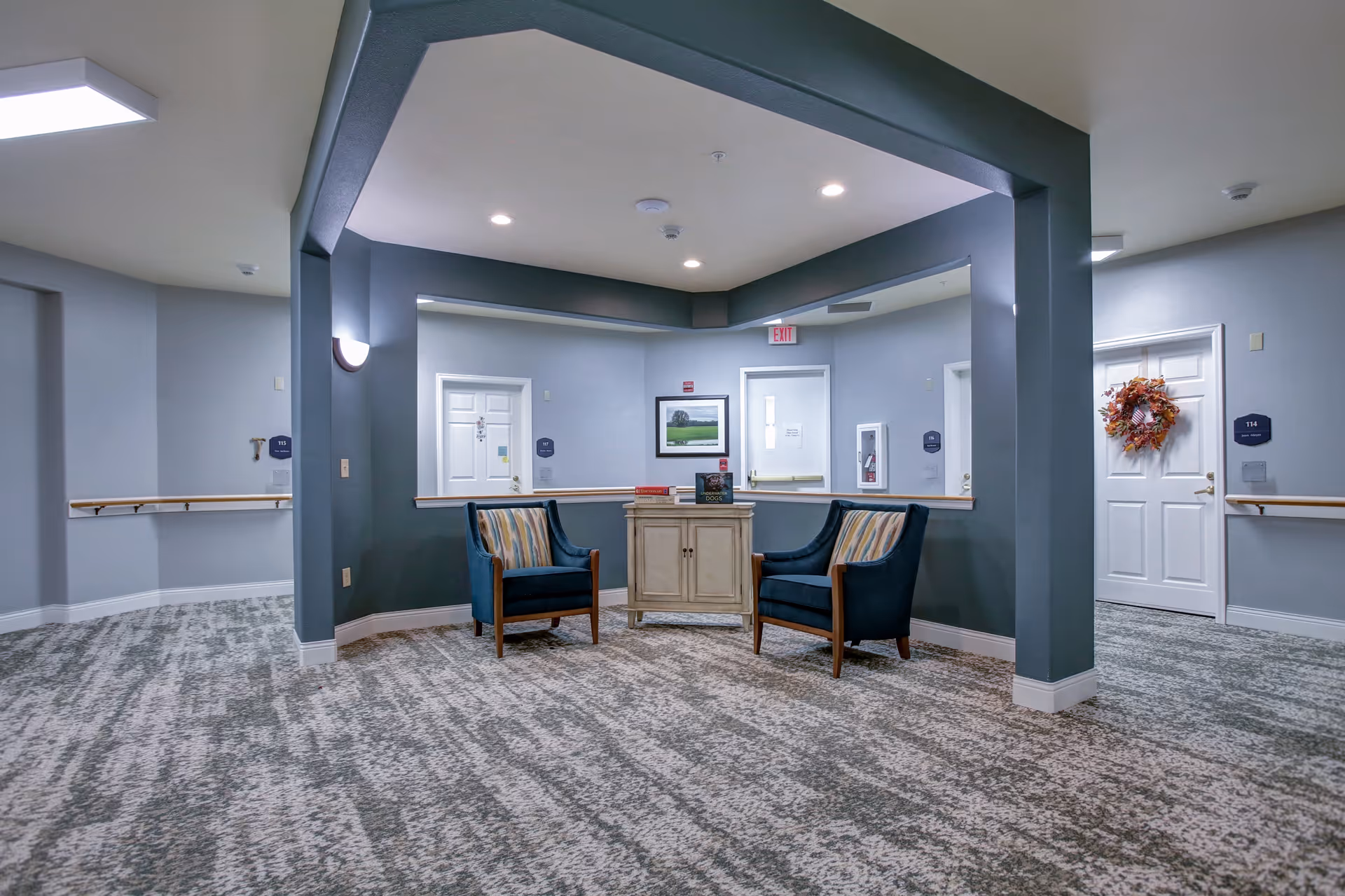 A quiet seating area in a senior living facility hallway with two blue armchairs and a small cabinet between them. The walls are painted gray with white doors labeled with room numbers. There is a framed picture on the wall and a wreath on one door. The carpet has a patterned design, and the ceiling has recessed lighting.