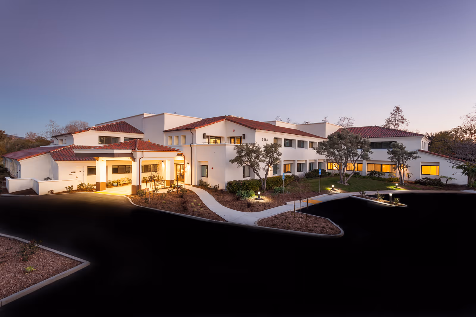 Exterior view of a two-story senior living facility building with white walls and red tile roof at dusk. The building is well-lit with warm lights, surrounded by landscaped greenery and a paved driveway and walkway leading to the entrance.