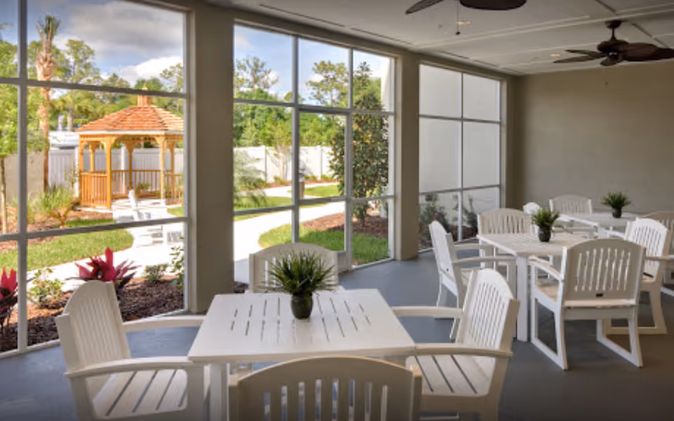 Sunroom seating area with white tables and chairs and small potted plants facing large windows that look out to a garden gazebo.
