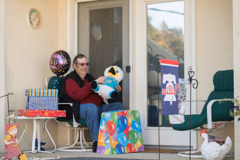 An elderly man sitting on a green cushioned chair on a porch holding a stuffed bird toy. There are several wrapped presents and a colorful gift bag with balloons on the ground next to him. A balloon with the number 80 is attached to the table beside him. The porch has a glass door with a Philadelphia Phillies flag hanging nearby and a white decorative chicken statue on the ground.