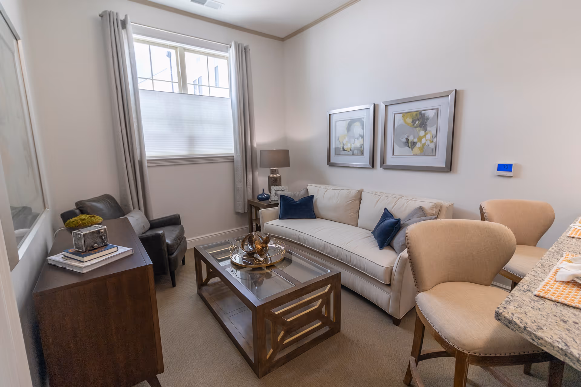 A bright furnished living room with a beige sofa, glass-top coffee table, armchairs, and a window with curtains.