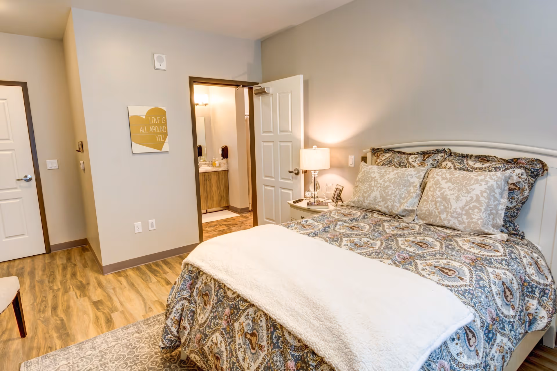 A cozy bedroom with a patterned bedspread and two decorative pillows on a white bed frame. A bedside table with a lamp and picture frame is next to the bed. The room has wood flooring and light-colored walls. An open door reveals a bathroom with a vanity and mirror. A wall decoration with the text 'LOVE IS ALL AROUND YOU' is visible.