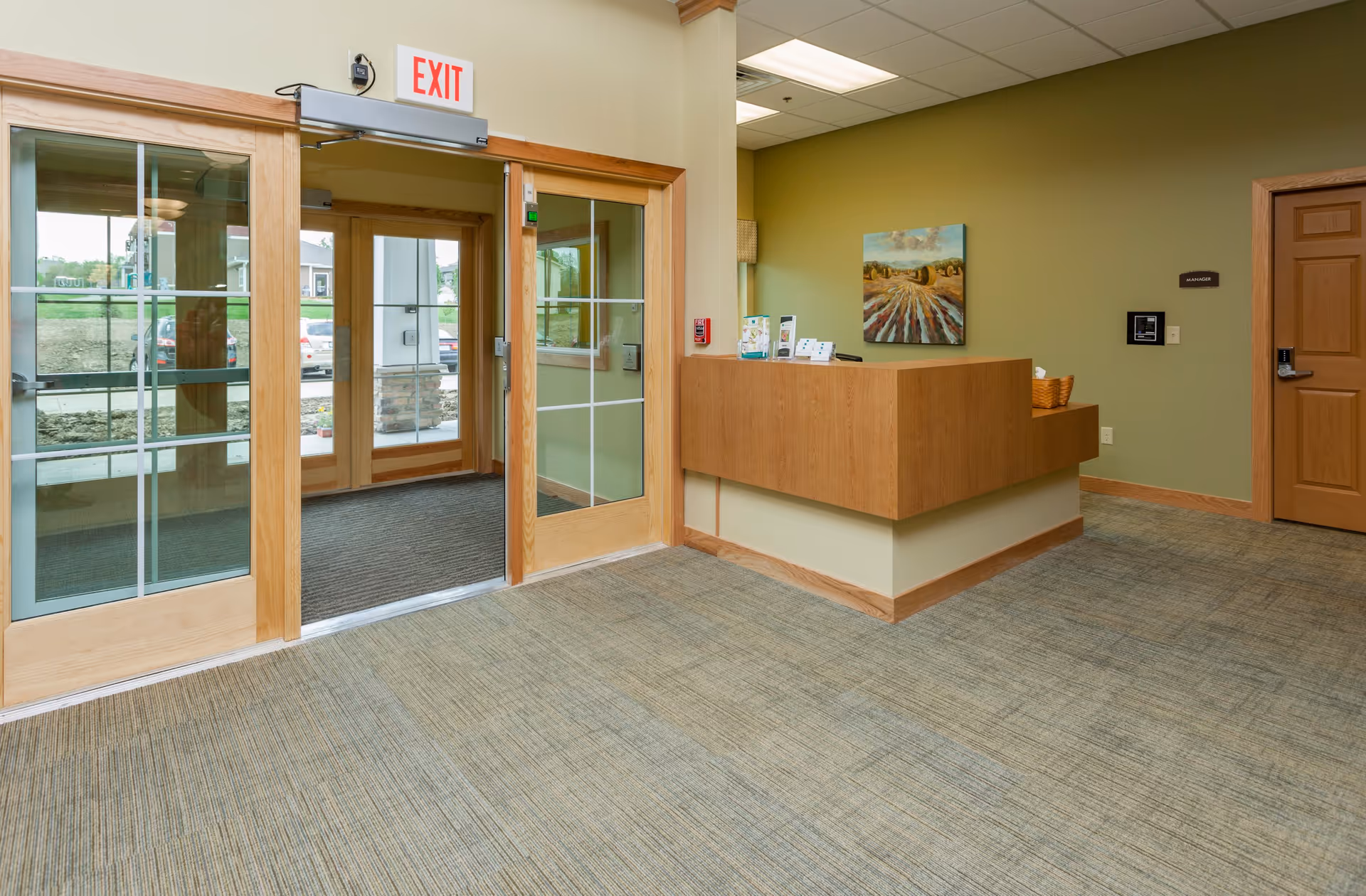 Reception area with a wooden front desk, green walls, a painting, and a glass double door exit leading outside. The floor is carpeted and there is a wooden door to the right of the desk.
