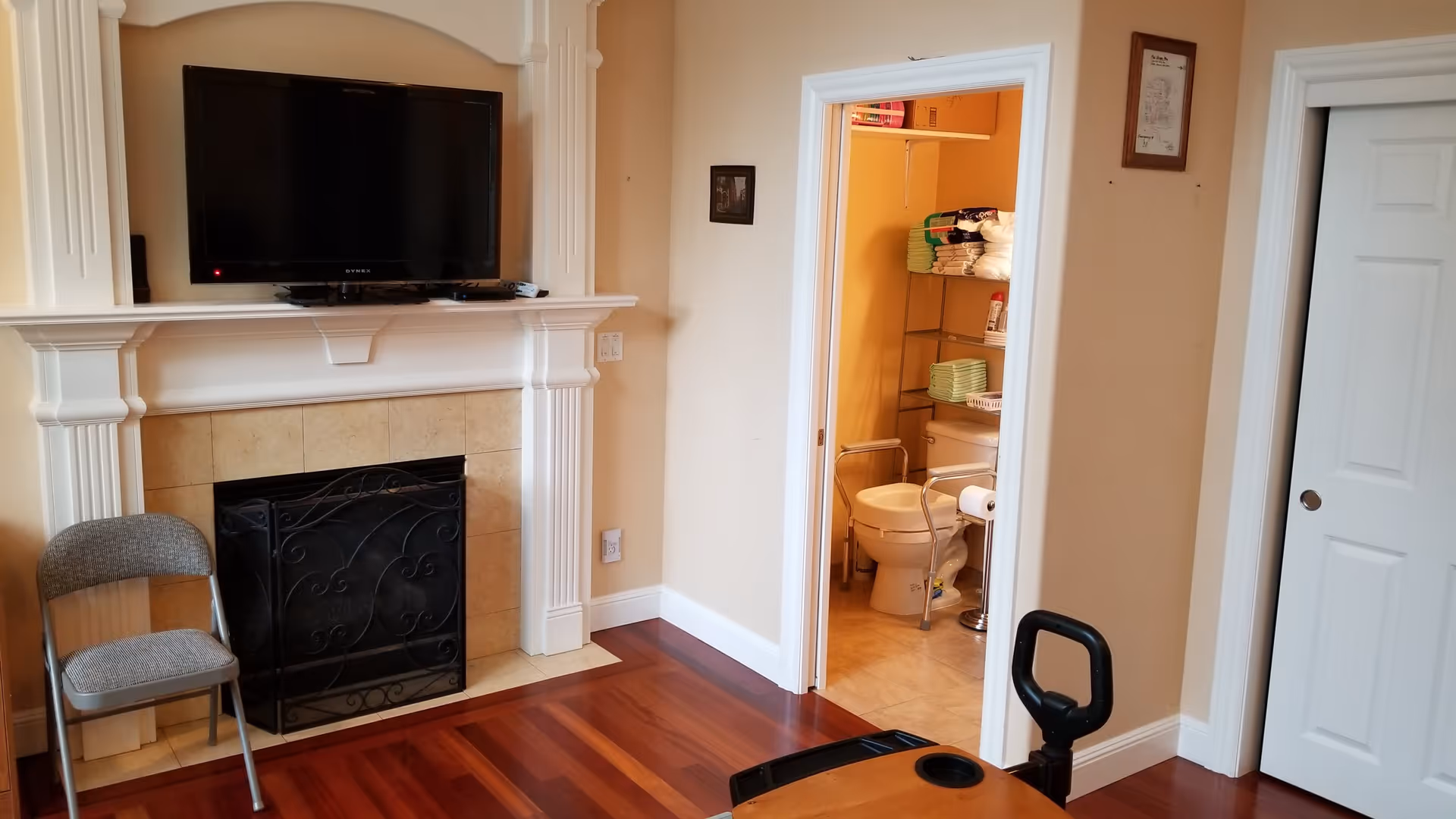 Interior view of a room with wooden flooring, a fireplace with a mounted flat-screen TV above it, a gray folding chair to the left of the fireplace, and an open door revealing a bathroom with a toilet, grab bars, and shelves with towels and supplies.
