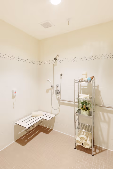 Accessible tiled shower room with a wall-mounted fold-down bench, handheld showerhead, grab bar, and a metal shelving unit holding towels and plants.