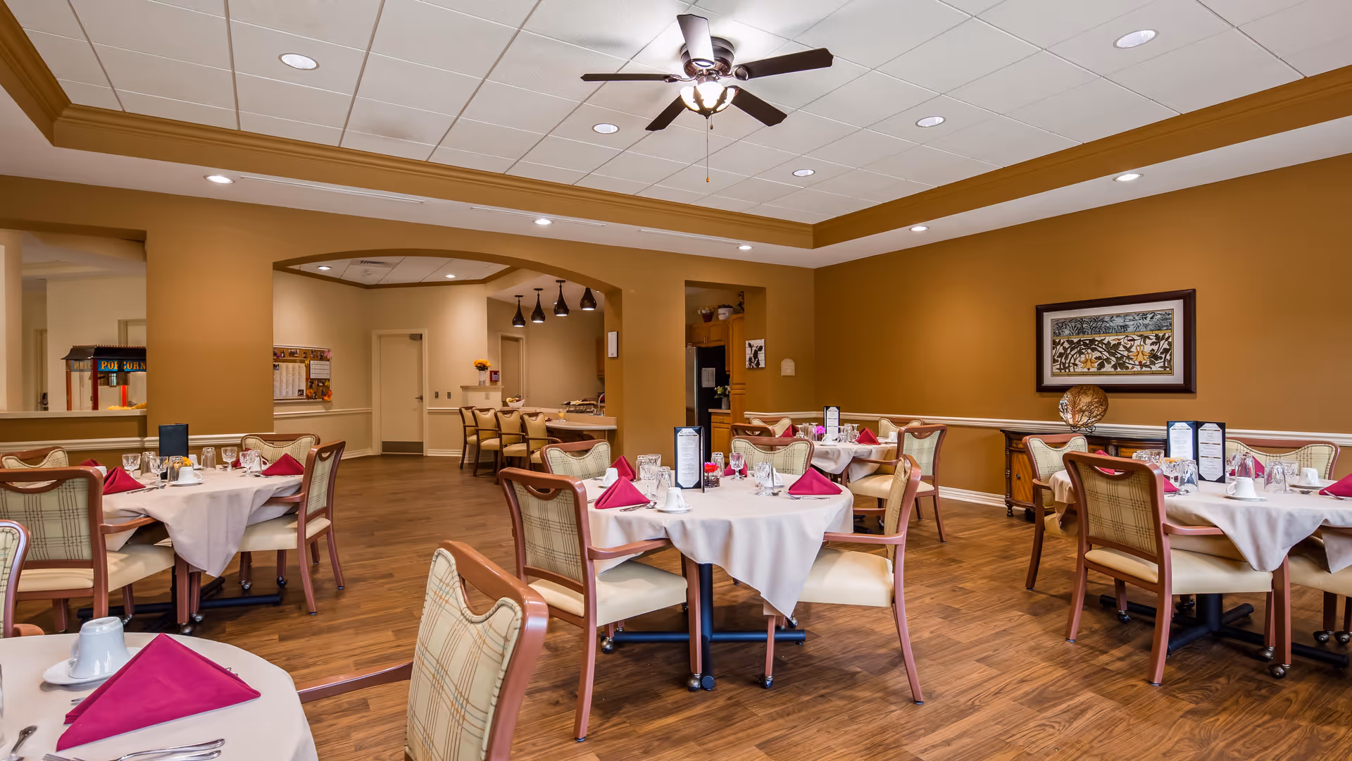 A dining room in a senior living facility with round tables covered in white tablecloths, each set with glasses, plates, silverware, and folded burgundy napkins. The room has wooden flooring, beige walls, and a ceiling fan with lights. There is a framed artwork on the wall and a sideboard with decorative items.