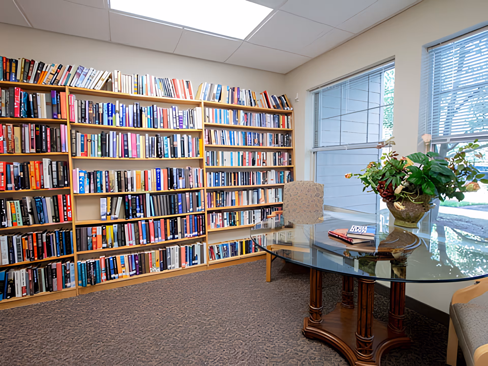 A cozy reading room with a large wooden bookshelf filled with books along the wall. There is a glass-top table with a decorative plant and a couple of books on it. Two windows with blinds allow natural light to brighten the room.