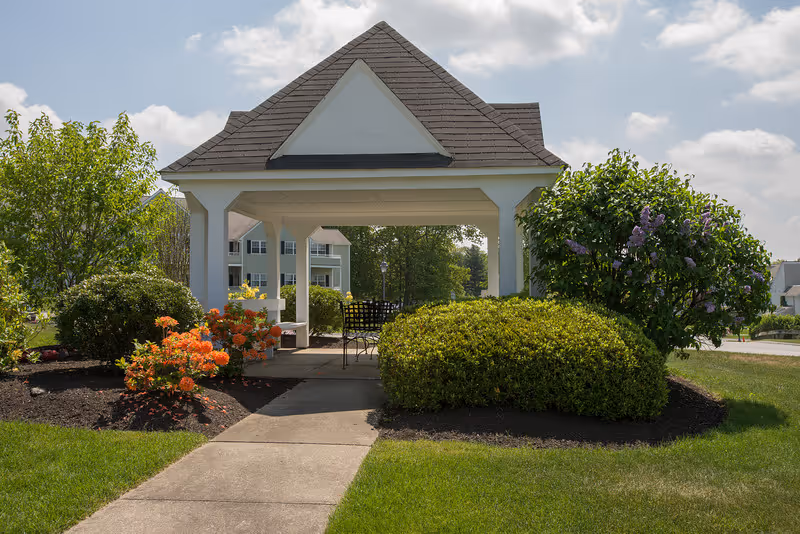 A small outdoor pavilion with a peaked roof and white pillars surrounded by green bushes and flowering plants. There is a concrete walkway leading to the pavilion, which contains a black metal chair and table set. Residential buildings and trees are visible in the background under a partly cloudy sky.