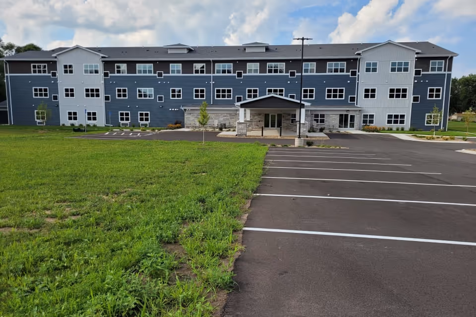 Front exterior view of a three-story residential building with blue and white siding, multiple windows, a covered entrance, a parking lot with marked spaces, and a grassy area in the foreground under a partly cloudy sky.