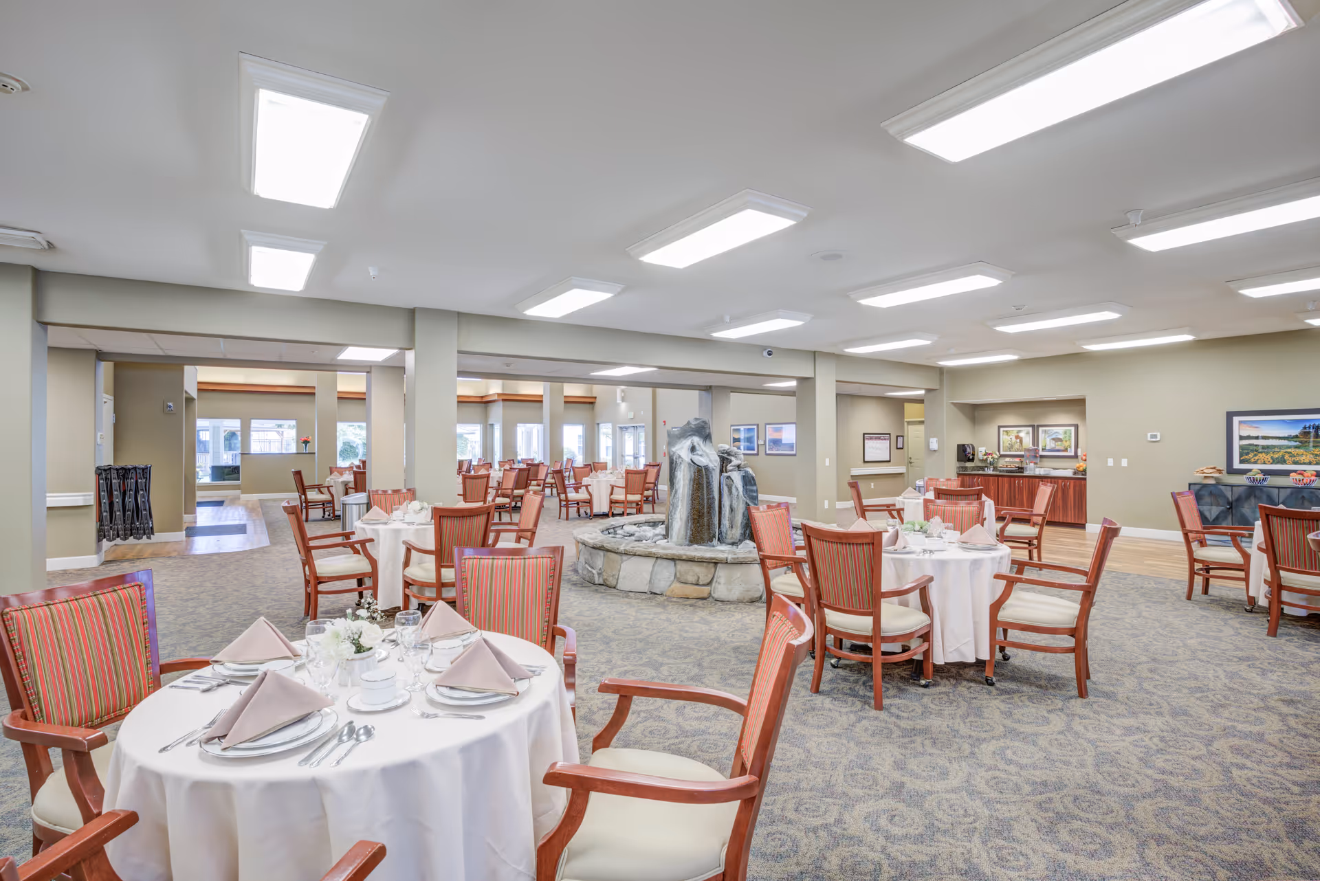 A spacious dining room in a senior living facility with round tables covered in white tablecloths, each set with plates, cups, napkins, and silverware. The room features wooden chairs with striped upholstery, a central stone water fountain, carpeted flooring, and bright overhead lighting. There are windows and framed pictures on the walls in the background.