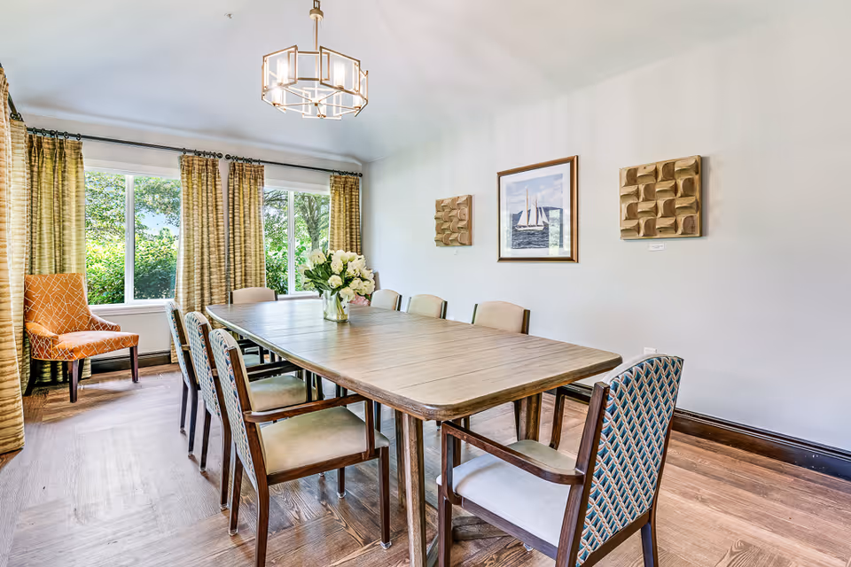 A bright dining room with a long wooden table surrounded by eight chairs, a vase with white flowers in the center, large windows with patterned curtains letting in natural light, a modern chandelier hanging from the ceiling, and three pieces of wall art including a framed picture of a sailboat.