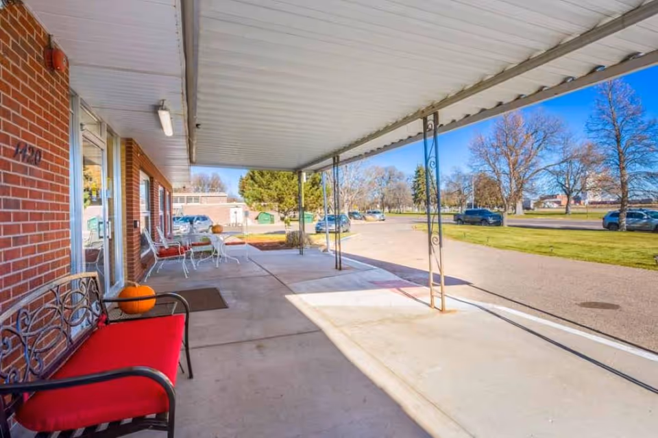 Covered outdoor patio area with a red cushioned bench and a small table with chairs. There are pumpkins on the bench and table. The patio is adjacent to a brick building with glass doors and windows. In the background, there is a parking lot with several cars and trees with no leaves under a clear blue sky.