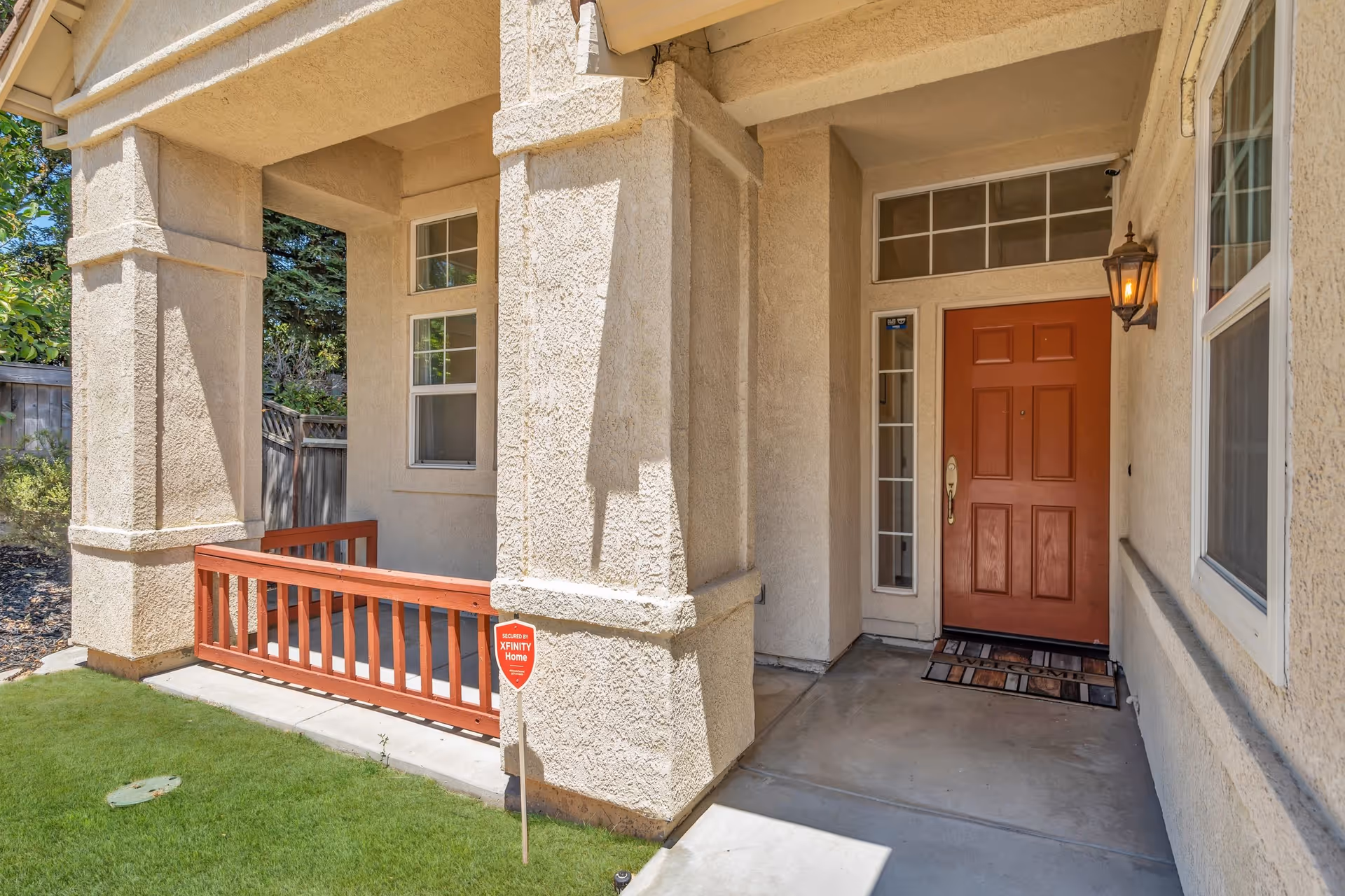 Front entrance of a stucco house showing a covered porch, orange door, columns, small red railing and an XFINITY Home security sign.