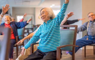 A group of elderly individuals seated on chairs in a room, participating in a seated exercise or stretching activity with their arms raised and smiling.